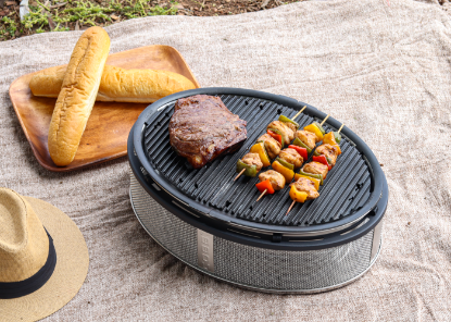 Picnic setting with the Cobb Supreme portable charcoal cooker grilling steak and kebabs on the Cobb griddle